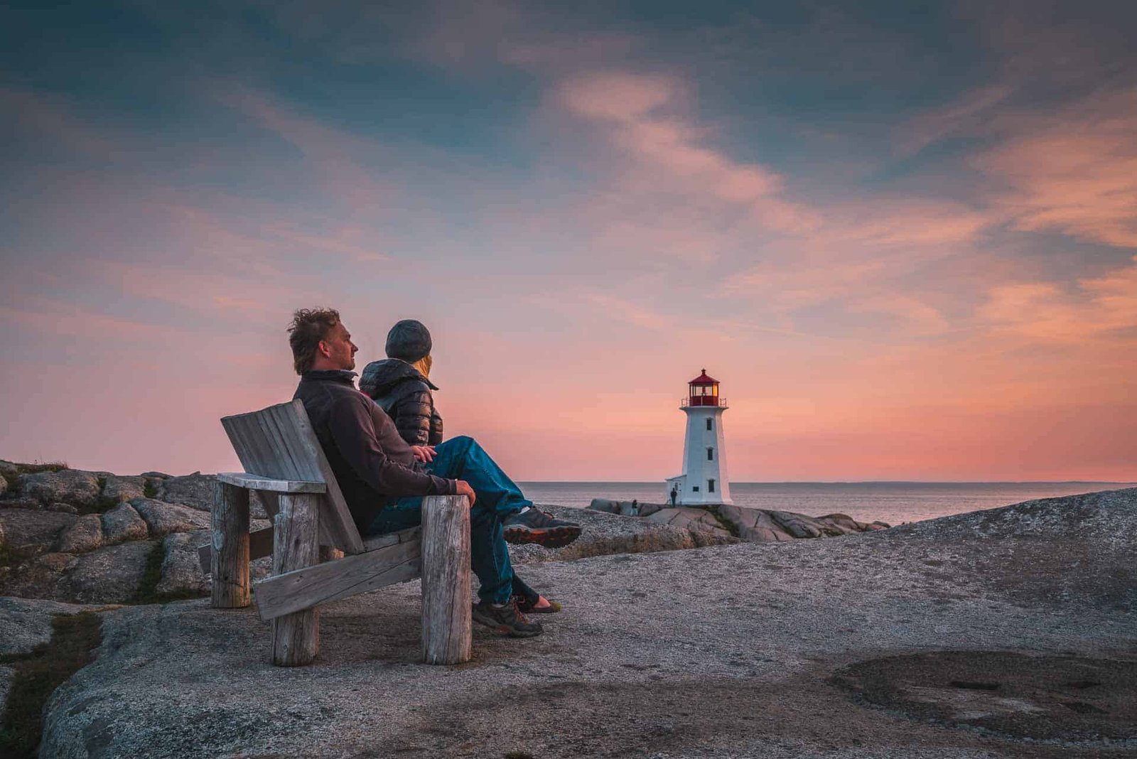 Dave and Deb of The Planet D at he iconic Peggy's Cove lighthouse at sunset, with waves crashing on the granite rocks in Nova Scotia.